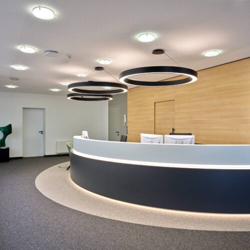 Banco reception curvo bicolore con LED integrato, parete boiserie rovere e tre anelli luminosi concentrici al soffitto.