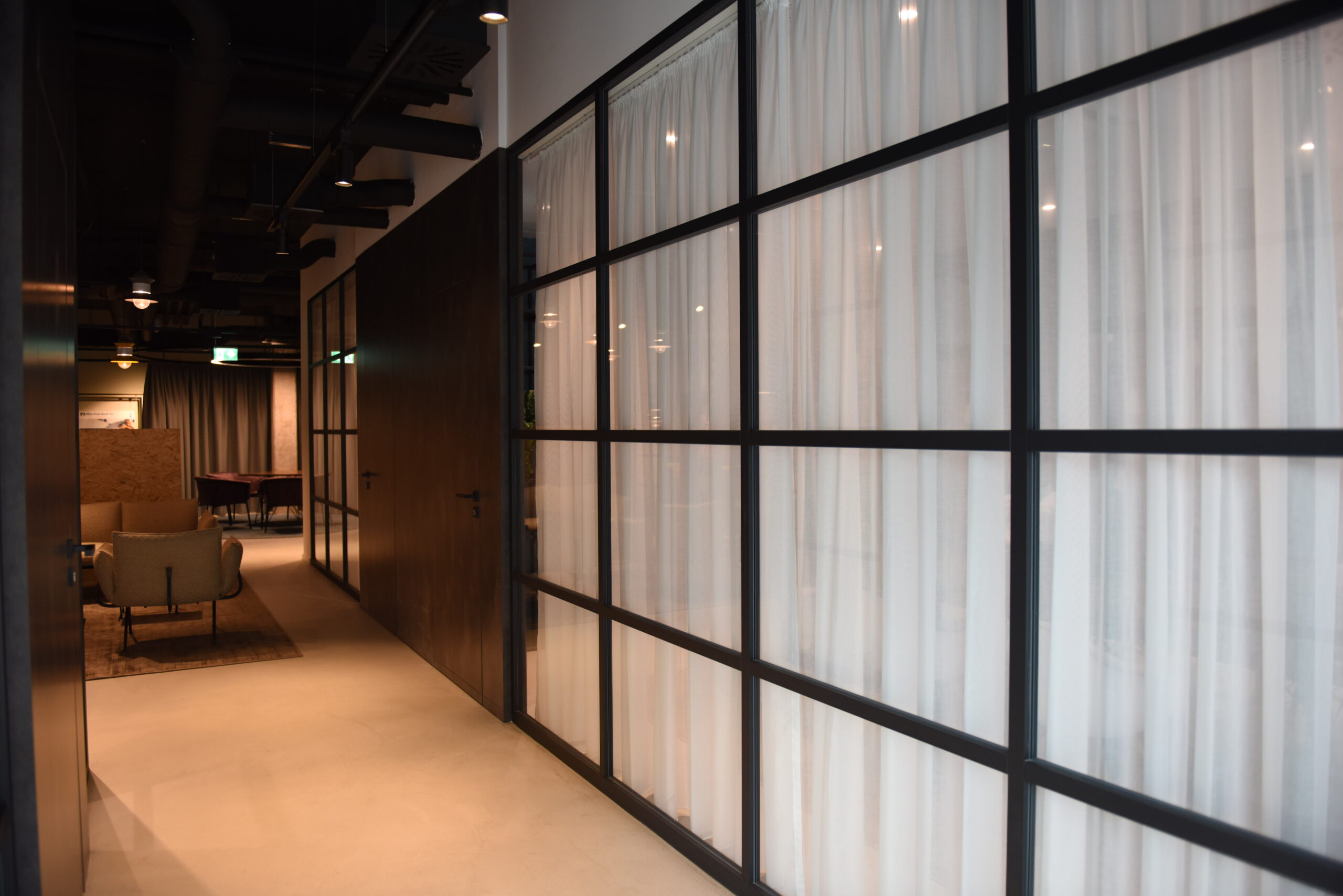 Corridoio minimal con pavimento in microcemento e parete vetrata a griglia nera con tende bianche.