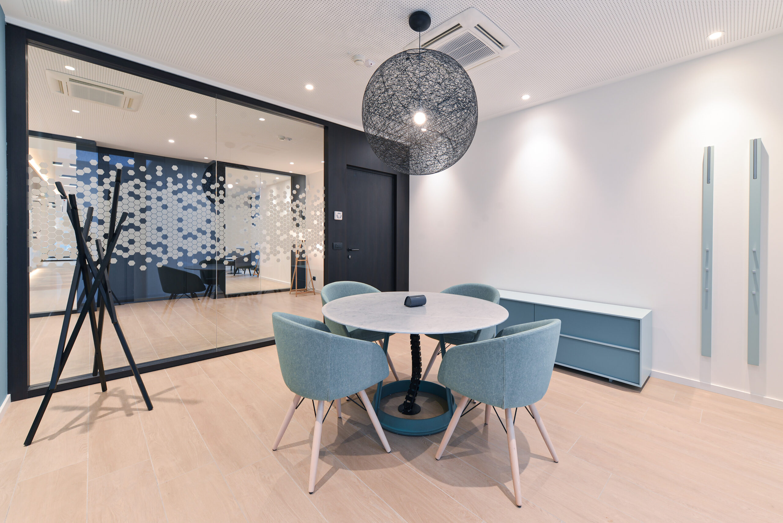 meeting informale Tavolo rotondo in pietra con quattro poltroncine verde salvia, lampada a sospensione sferica in rete metallica e parete vetrata serigrafata con motivo esagonale.