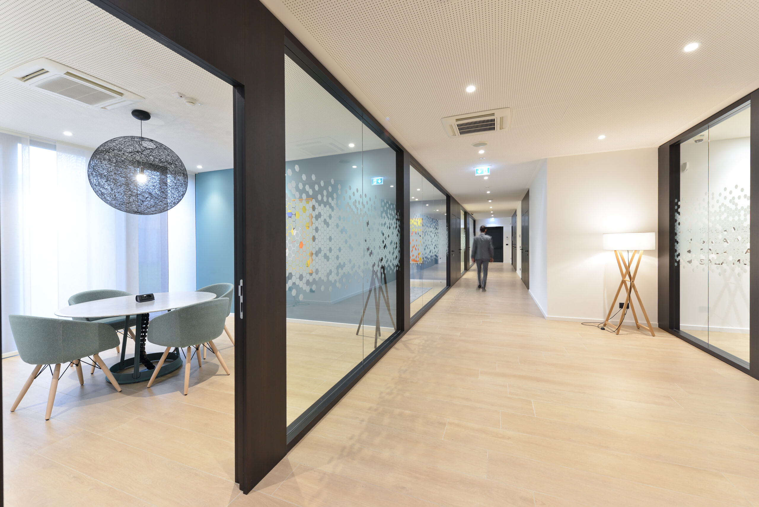 Lungo corridoio con pavimento in legno chiaro e uffici separati da pareti vetrate serigrafate; a sinistra area meeting, a destra lampada da terra treppiede.