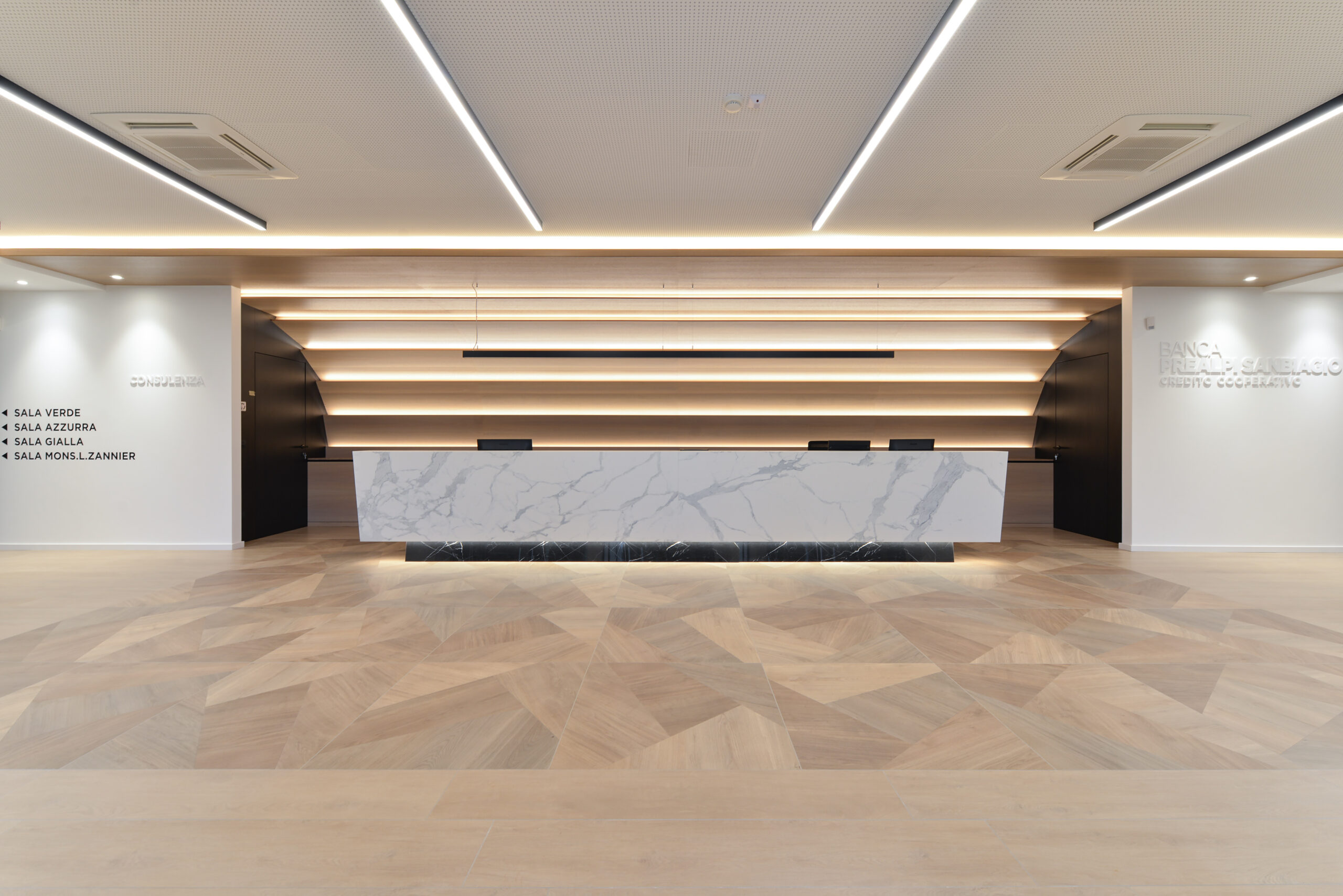 Banco accoglienza monolitico in marmo bianco davanti a parete retro-illuminata a listelli orizzontali in legno chiaro.