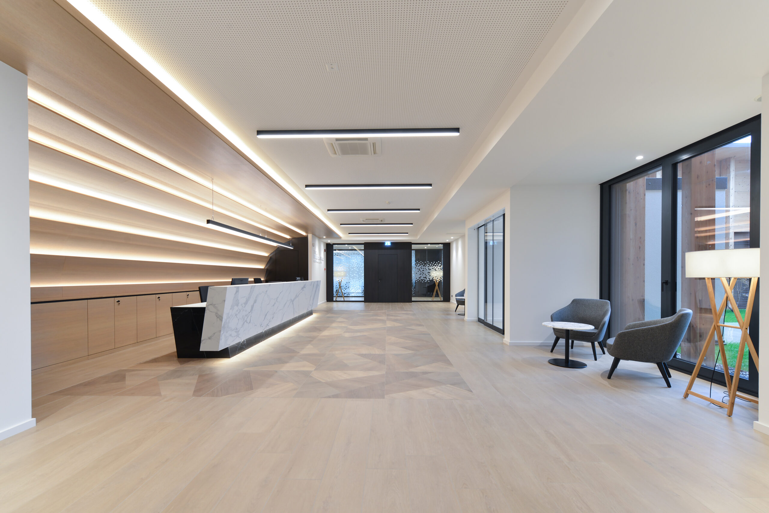 Ampio atrio con reception in marmo, pavimento geometrico in essenza rovere e zona attesa con poltrone grigie e lampada treppiede.