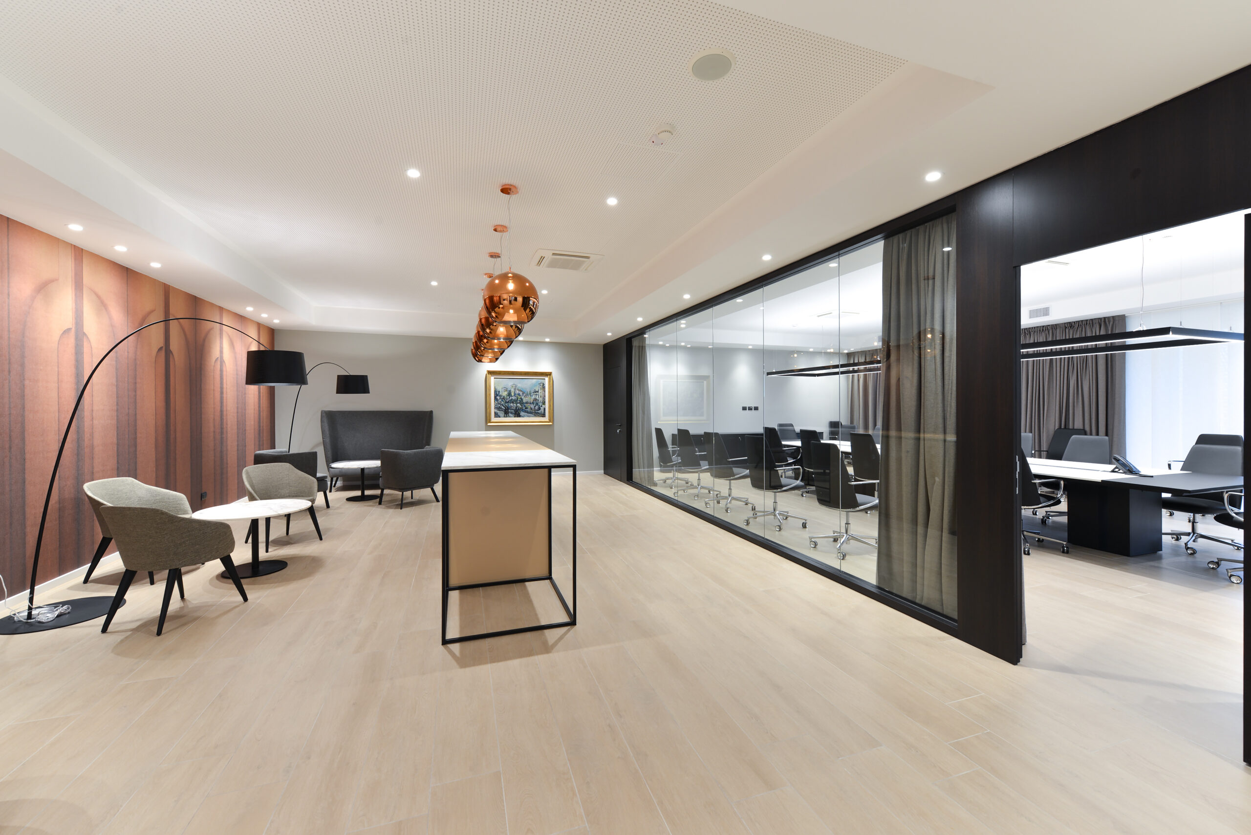 Vista d’insieme su lounge con poltroncine, tavoli laterali e lampade a sospensione in rame; al centro bancone alto, a destra sala riunioni vetrata con tavolo direzionale.