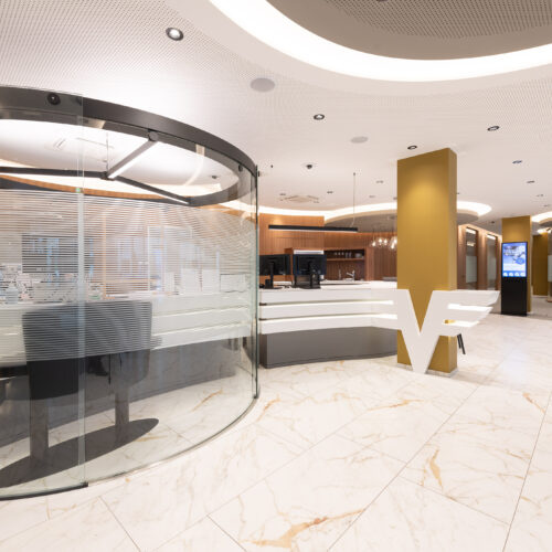 Zona reception con desk circolare in vetro serigrafato, logo “V” in primo piano, pavimento in gres effetto marmo e colonne rivestite in ottone.