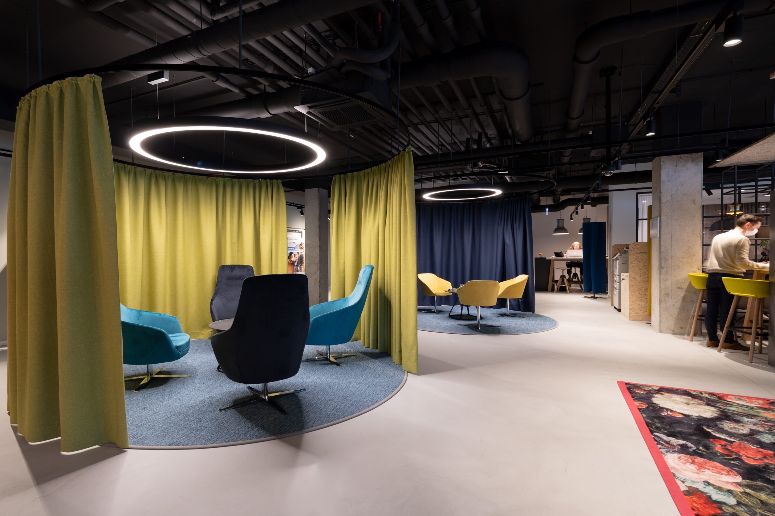 Postazioni di conversazione circolari delimitate da tende verdi e blu con poltrone imbottite e lampade ad anello LED.