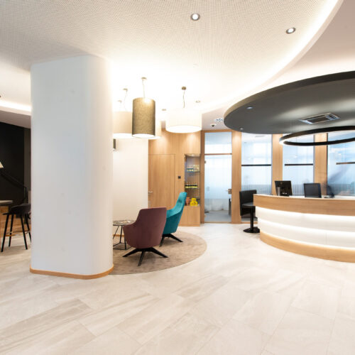 Atrio luminoso con desk ellittico retro-illuminato, soffitto acustico nero a cupola con anello LED, colonne bianche e poltrone colorate su pavimento in gres chiaro.