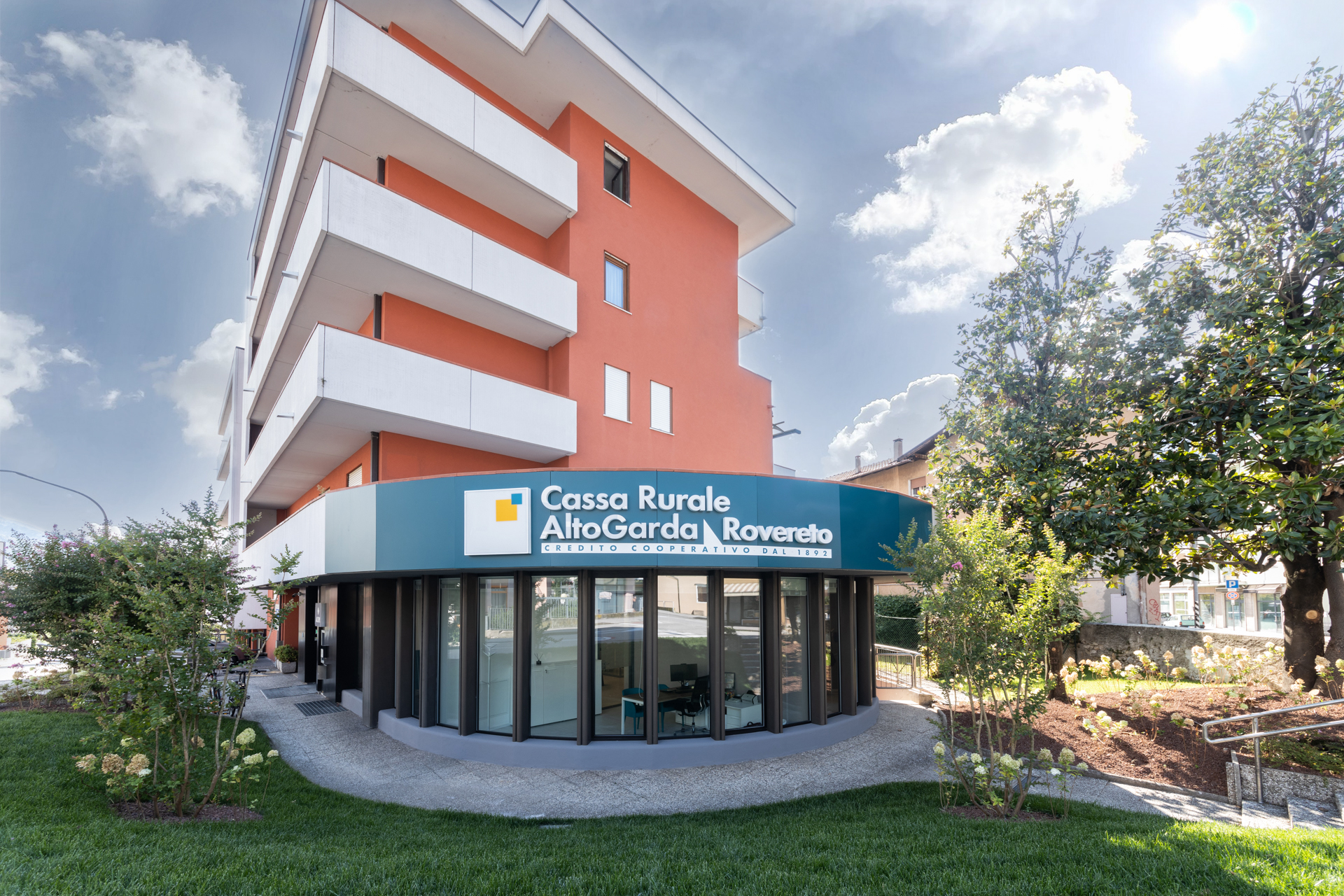 Facciata filiale curva al piano terra di palazzina arancione con balconi bianchi, insegna blu Cassa Rurale AltoGarda Rovereto e giardino anteriore curato.