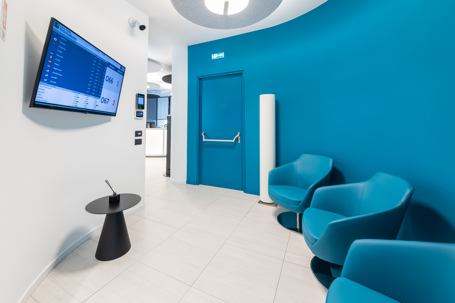 Angolo attesa: tre poltrone blu petrolio, tavolino nero e lampade fonoassorbenti circolari a soffitto.