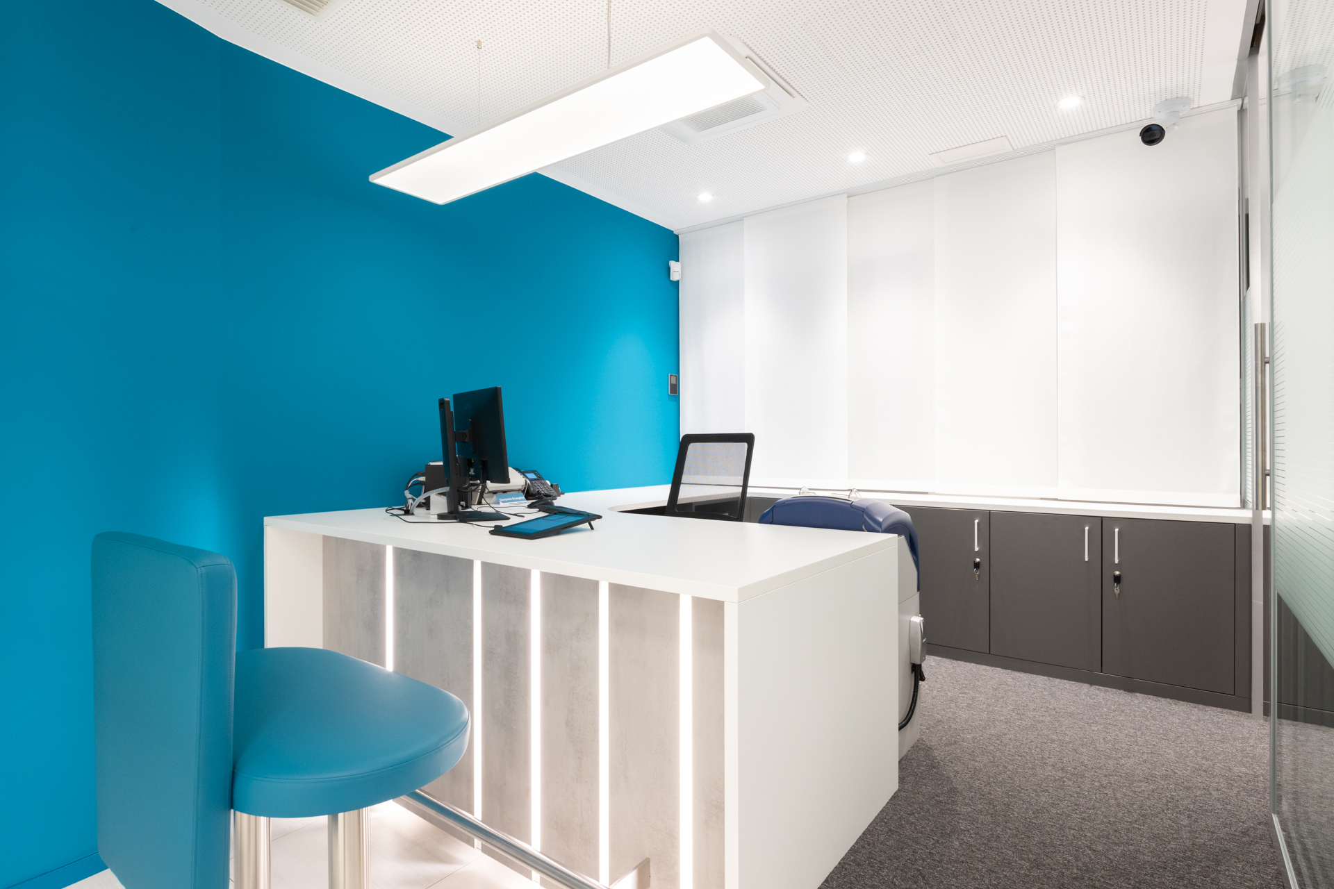Postazione operativa con bancone bianco retro-illuminato, sgabello imbottito blu, parete teal a contrasto e grandi tende bianche filtranti alle finestre.