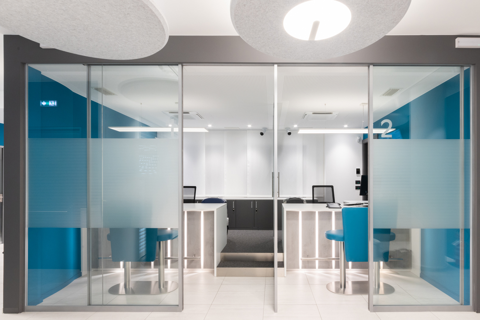 Box consulenza number-2 vetrato con vetrofanie satinate, due desk retro-illuminati, sedute blu e pannelli acustici circolari a soffitto.