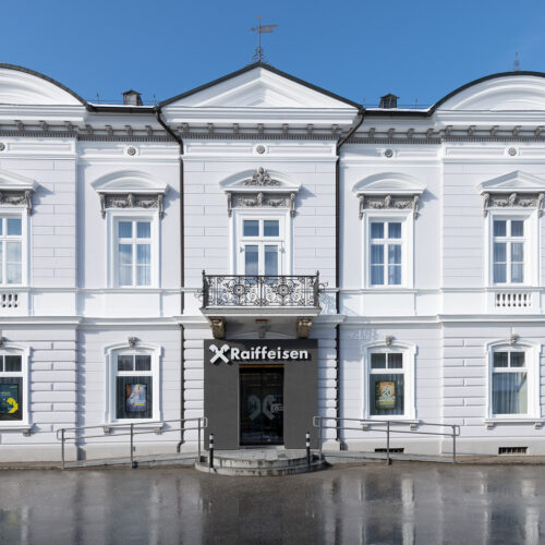 Prospetto frontale di edificio neoclassico tinteggiato di bianco con cornici e lesene, insegna Raiffeisen sopra ingresso centrale.