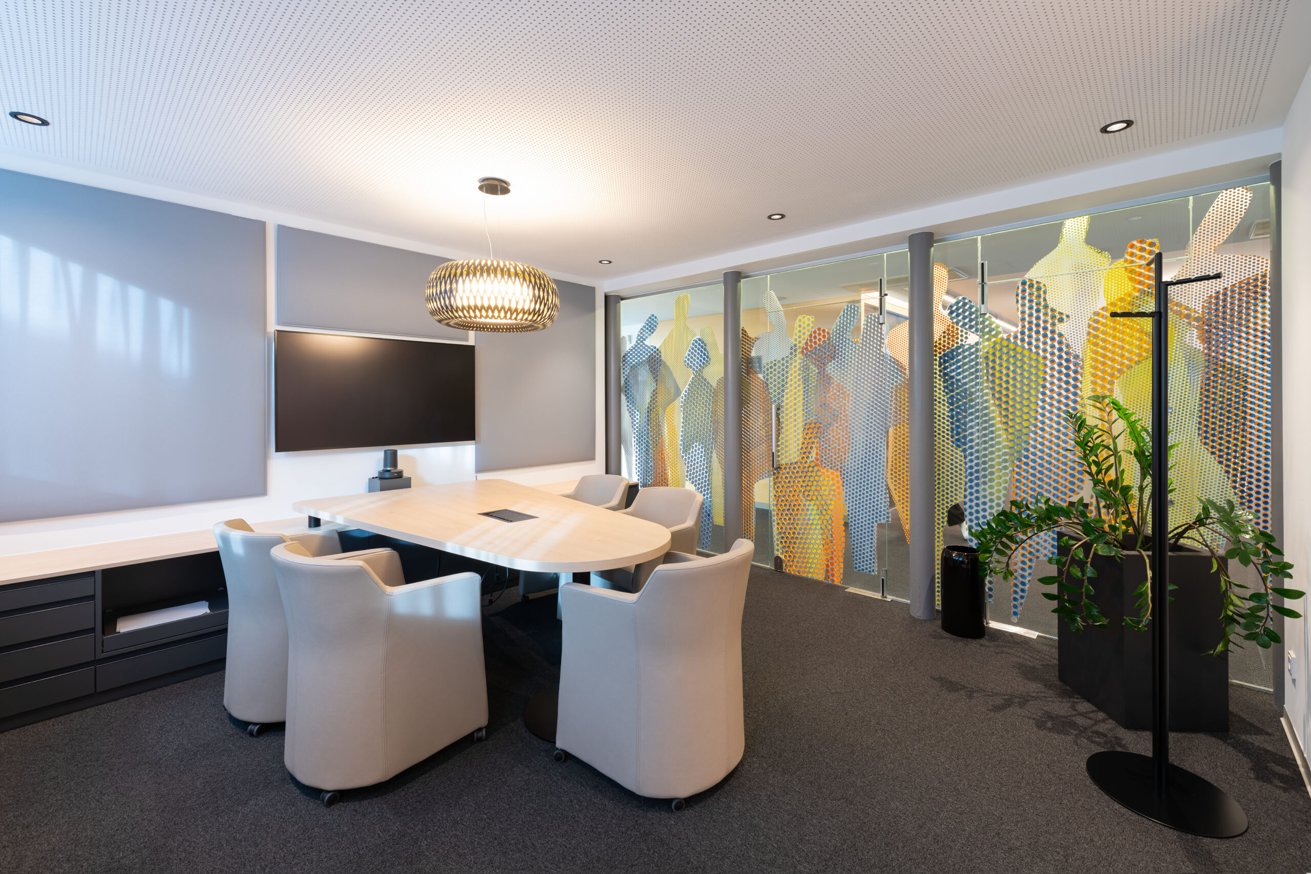 Sala riunioni con tavolo ovale in legno chiaro, poltrone imbottite, lampadario intrecciato e parete vetrata con grafiche a silhouette nella Raiffeisen Regionalbank Schwaz-Wattens