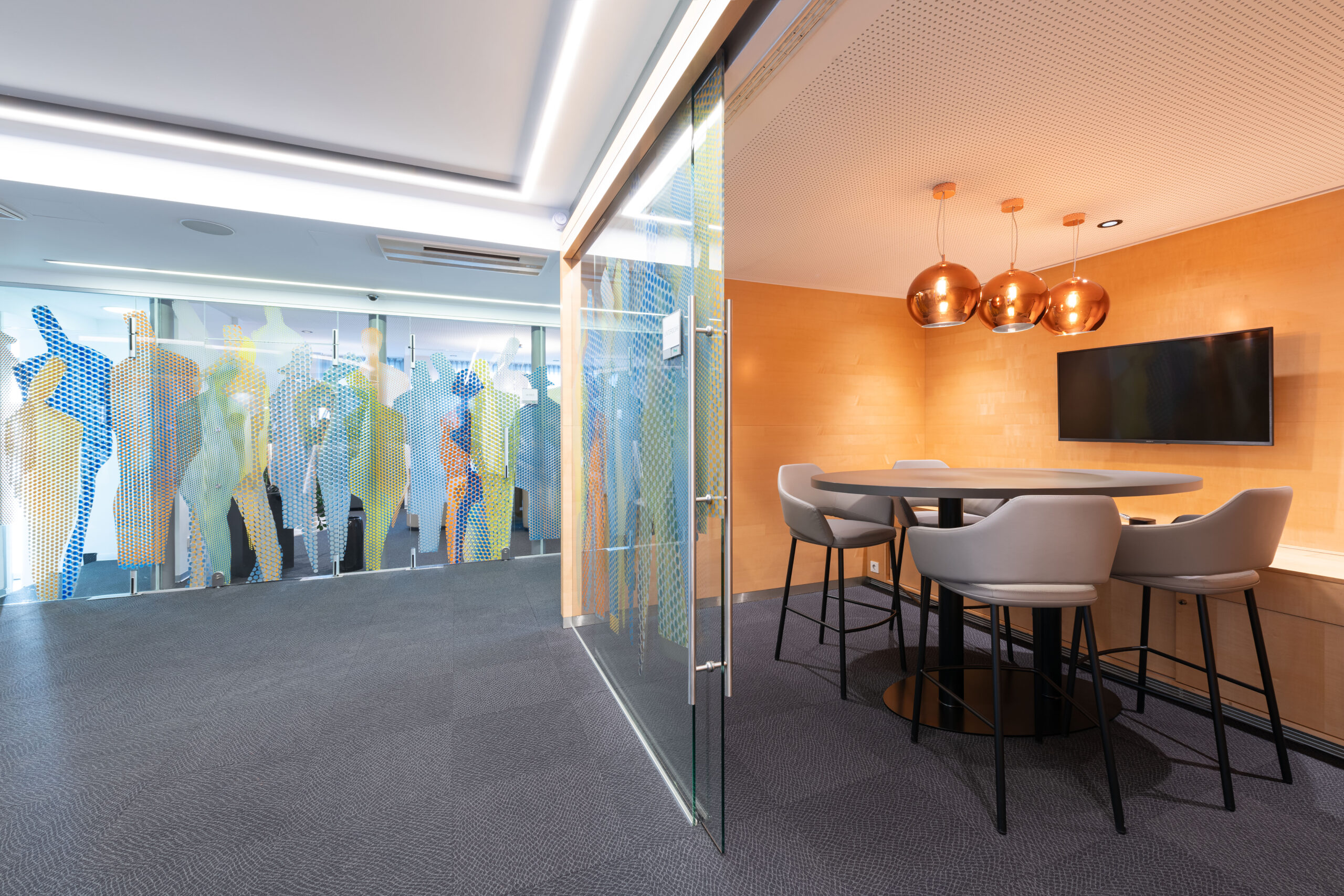 Piccolo meeting corner rivestito in legno con tavolo alto rotondo, sgabelli imbottiti e lampade a sospensione in rame, adiacente a corridoio vetrato decorato nella Raiffeisen Regionalbank Schwaz-Wattens