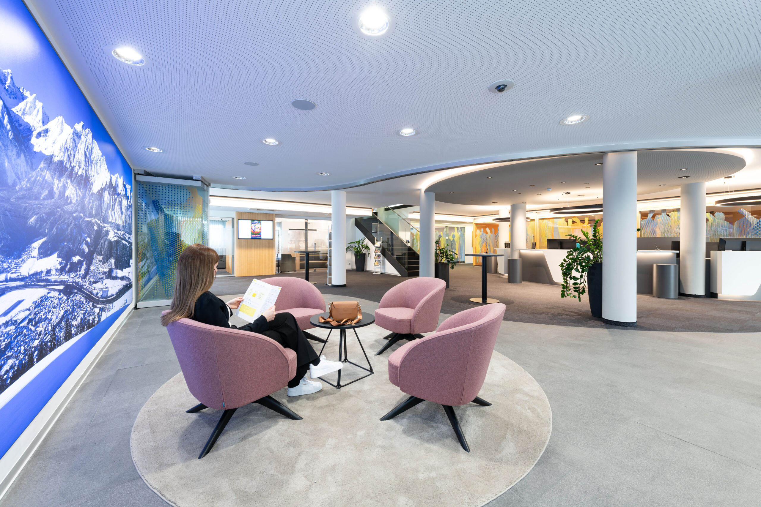 Area attesa con poltrone rosa e tavolini, vista sull’atrio circolare e sulle postazioni reception della Raiffeisen Regionalbank Schwaz-Wattens.