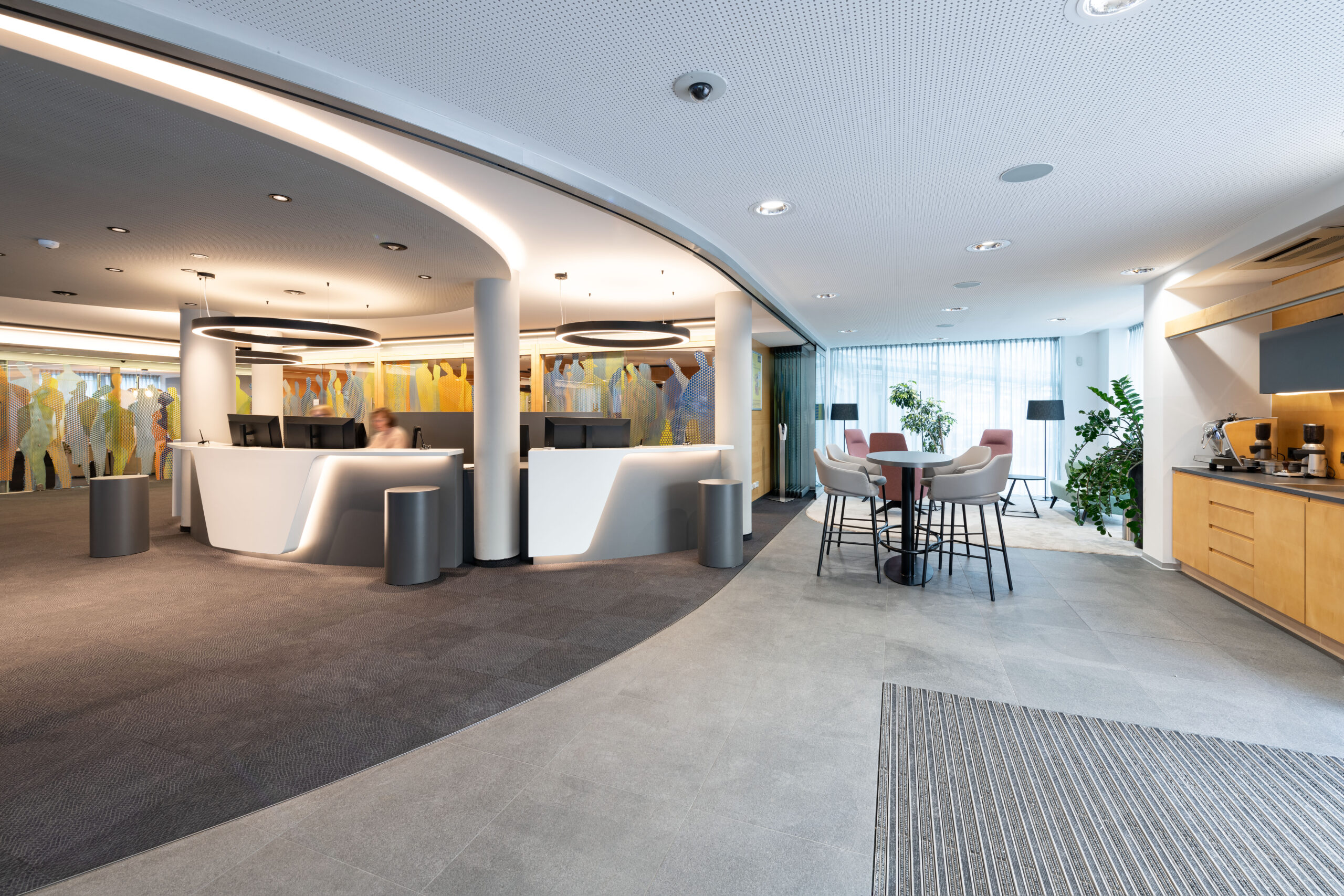 Vista dell’area reception con banconi bianchi retro-illuminati, lampade a disco sospese, tavolo alto con sedie imbottite e corner caffè in legno negli interni della Raiffeisen Regionalbank Schwaz-Wattens progettati da Dreika.