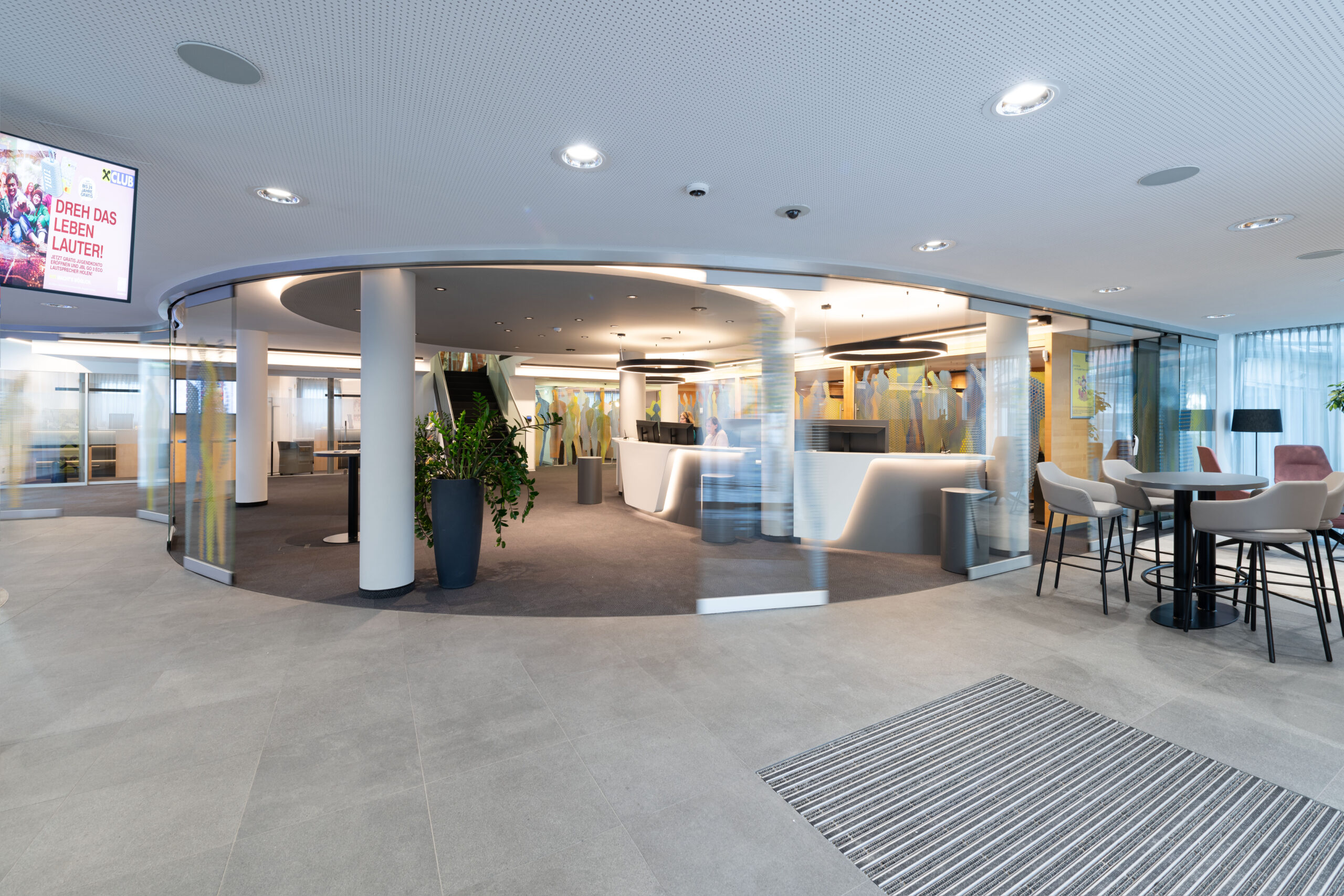 Atrio circolare con pareti curve in vetro serigrafato a silhouette colorate, pavimento in gres grigio e banconi reception luminosi della filiale Raiffeisen Regionalbank Schwaz-Wattens.