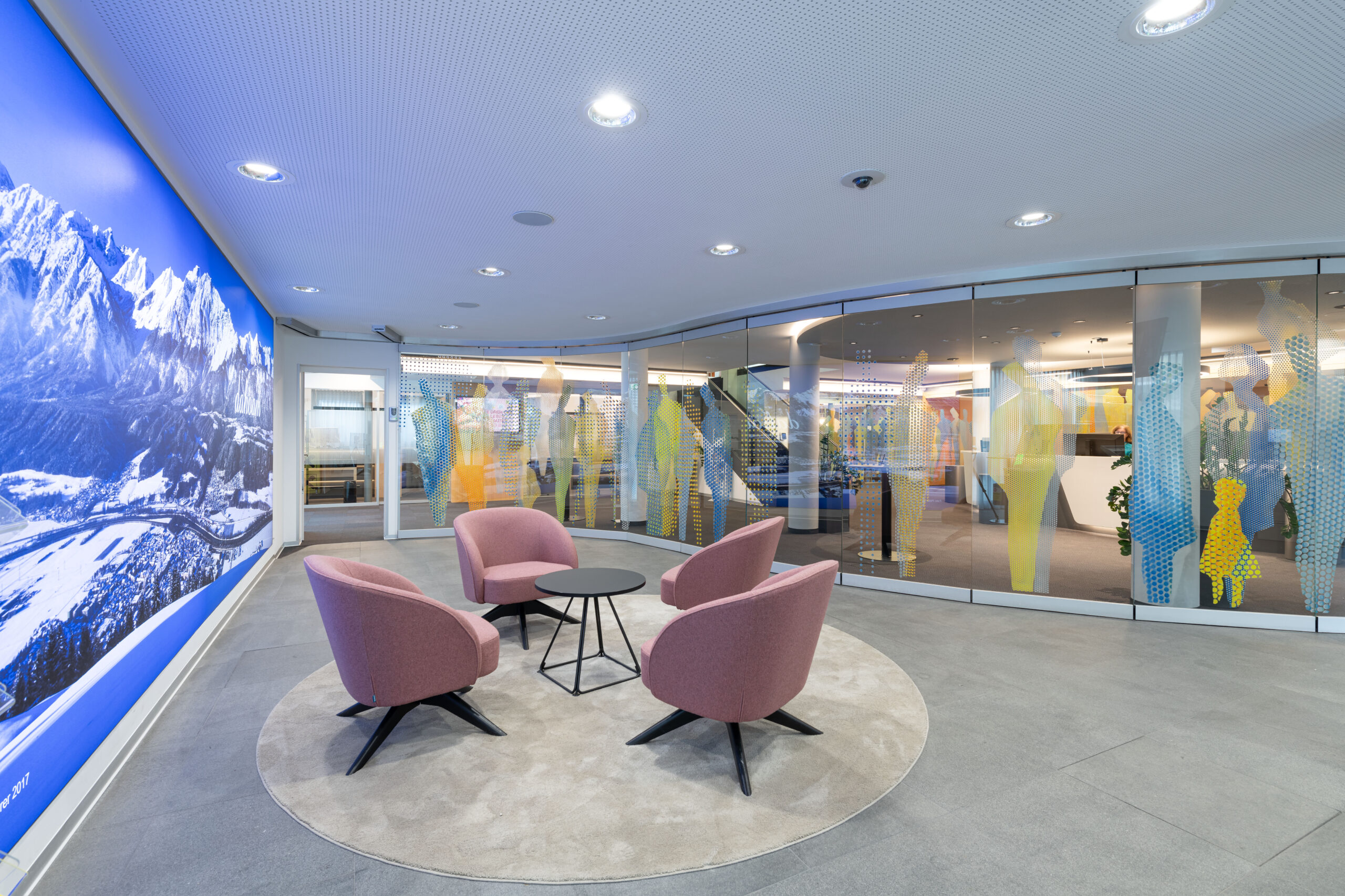 Zona lounge con quattro poltrone rosa su tappeto rotondo, maxi murale fotografico delle Alpi e divisorio vetrato illustrato negli interni della Raiffeisen Regionalbank Schwaz-Wattens.