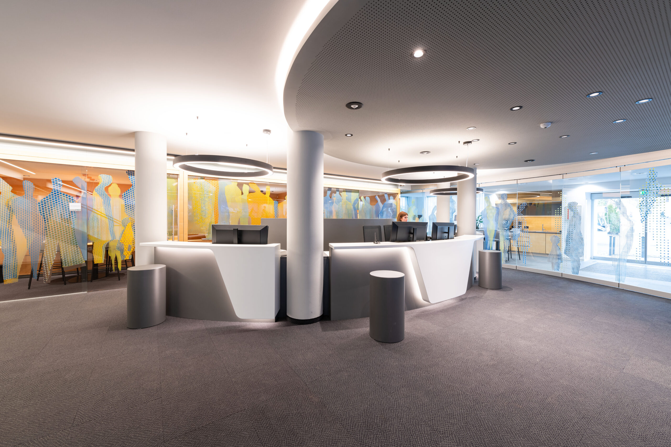 Banconi front office bianchi con illuminazione LED, soffitto microforato scuro e pareti artistiche colorate negli spazi aperti della Raiffeisen Schwaz-Wattens.