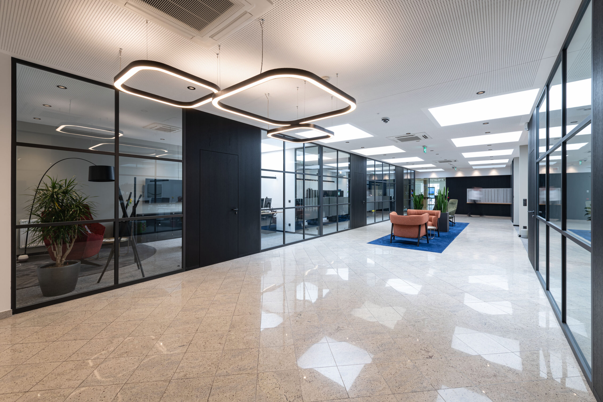 Spazio aperto con pavimento lucido, poltrone rosa su tappeto blu, illuminazione moderna e uffici a vetro.