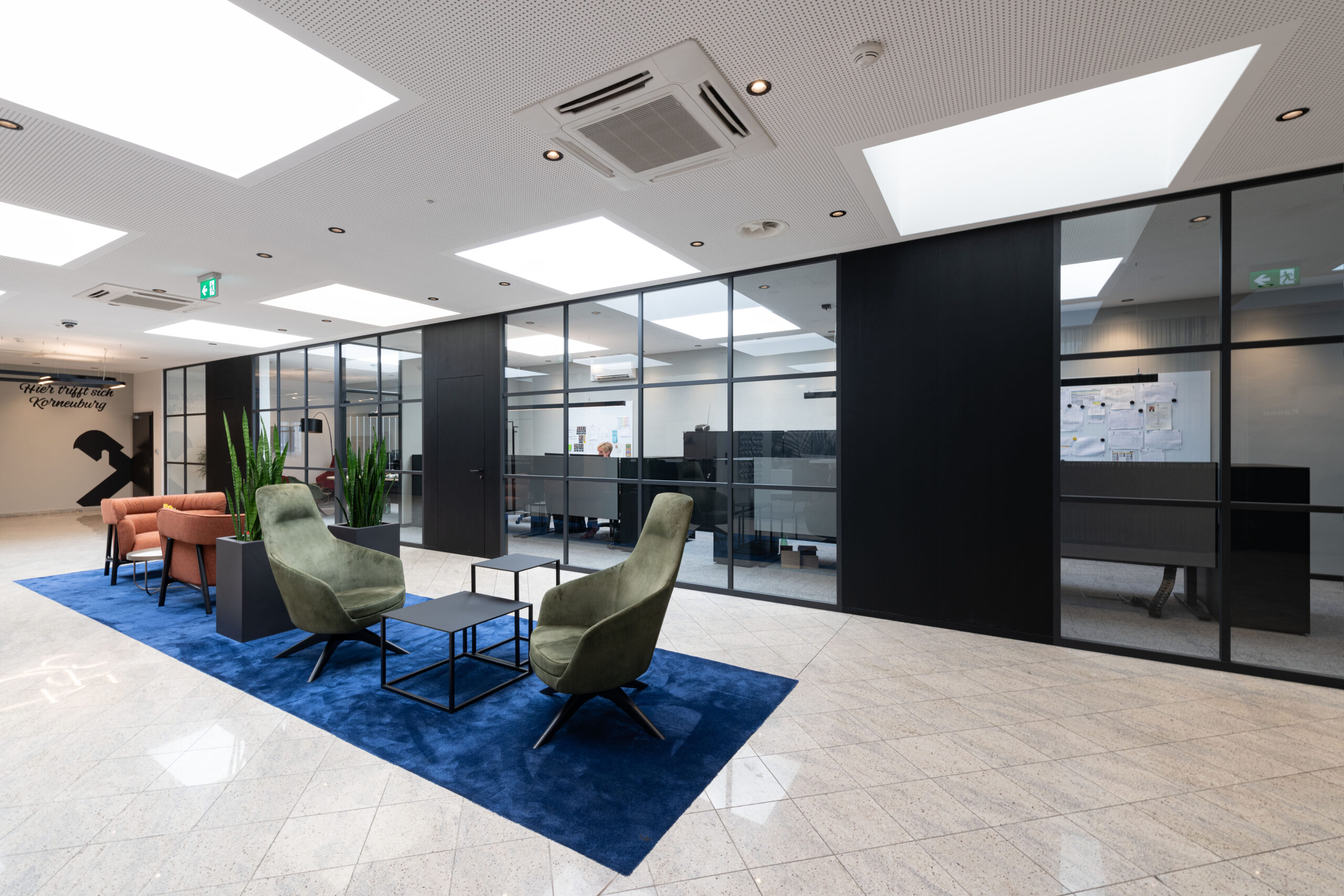 Zona attesa con poltrone verdi e arancioni, tappeto blu e pareti divisorie in vetro con vista su postazioni operative.