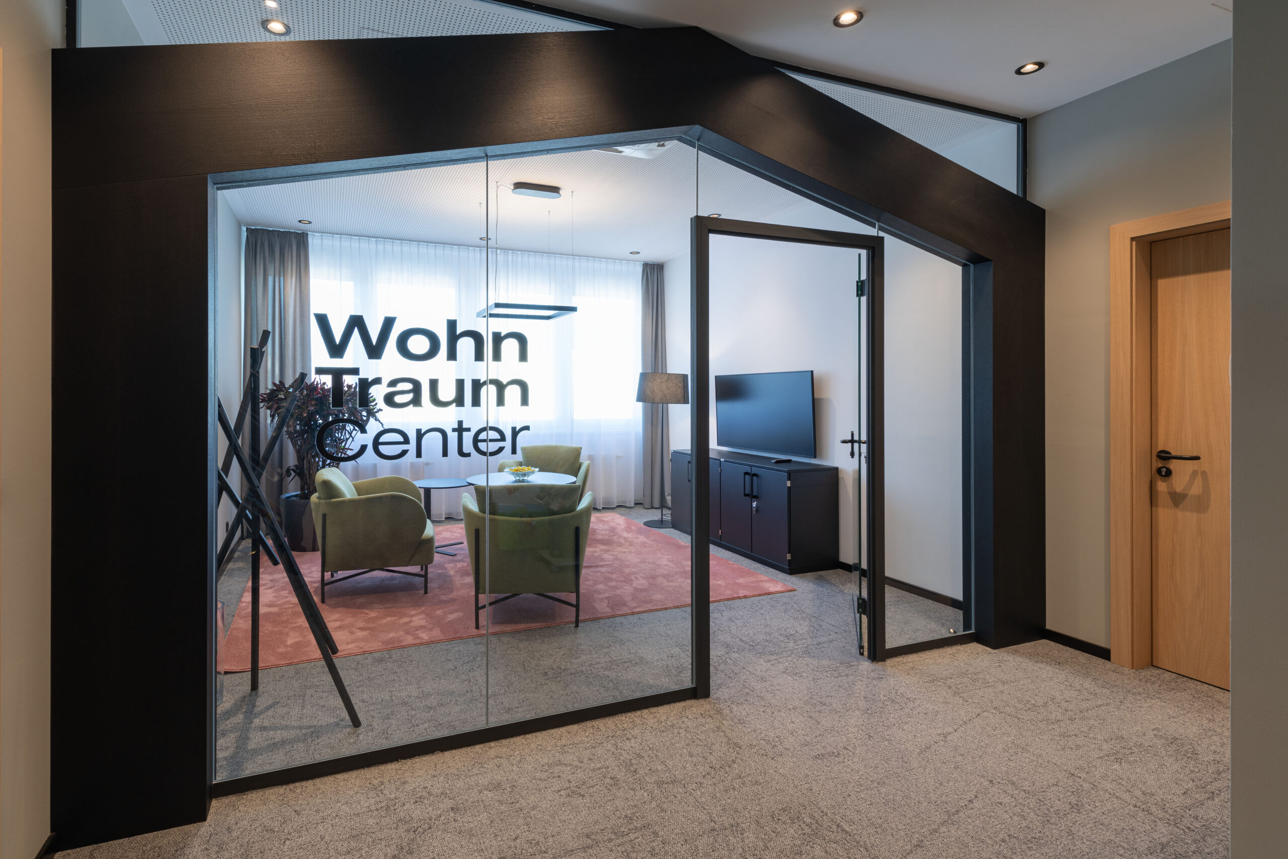 Ingresso vetrato con scritta “Wohn Traum Center” e area interna con tavolino, poltrone verdi e tappeto rosa.